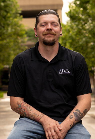 Man with tattoos and earrings sitting outdoors in front of greenery, wearing a black polo shirt with 'KDA' logo.