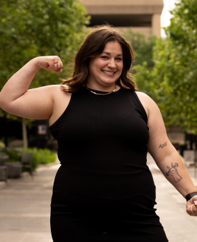 A woman wearing a black sleeveless top, smiling, and flexing her right arm outdoors in a park-like setting with trees and buildings in the background.