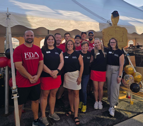 Group of nine people standing together outdoors under a white tent, smiling, some wearing red or black KDA shirts, with a large golden Oscar statue and gold, black, and white balloons in the background.