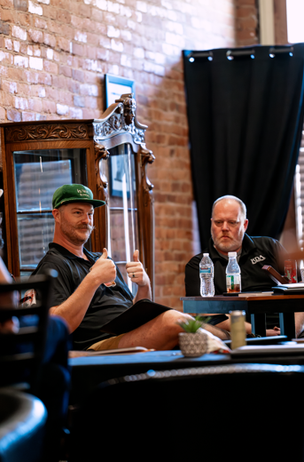 Two men sitting at a table in a room with brick walls, one giving a thumbs-up and the other looking down. There are water bottles and a laptop on the table, and a large mirror and black curtain in the background.