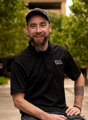 A smiling man with a beard, wearing a black cap and polo shirt, sitting outdoors with green trees in the background.