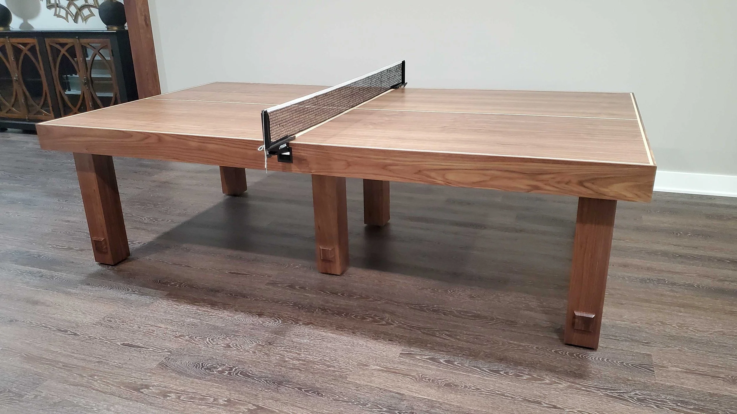 A wooden ping pong table with a net set up in a room with hardwood floors, white walls, and a decorative cabinet in the background.