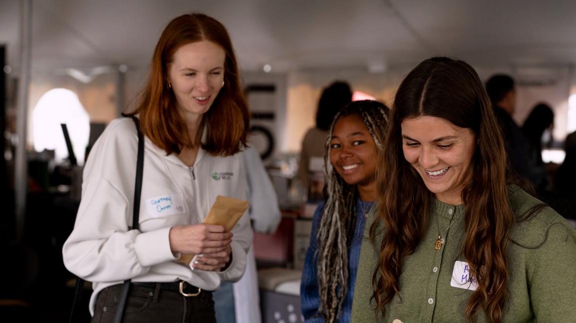 Three women at an indoor event, smiling and engaging in conversation. The woman on the left is holding a paper bag, wearing a white jacket and a name tag. The woman on the right has long brown hair, a green shirt, and a name tag, and is looking down.