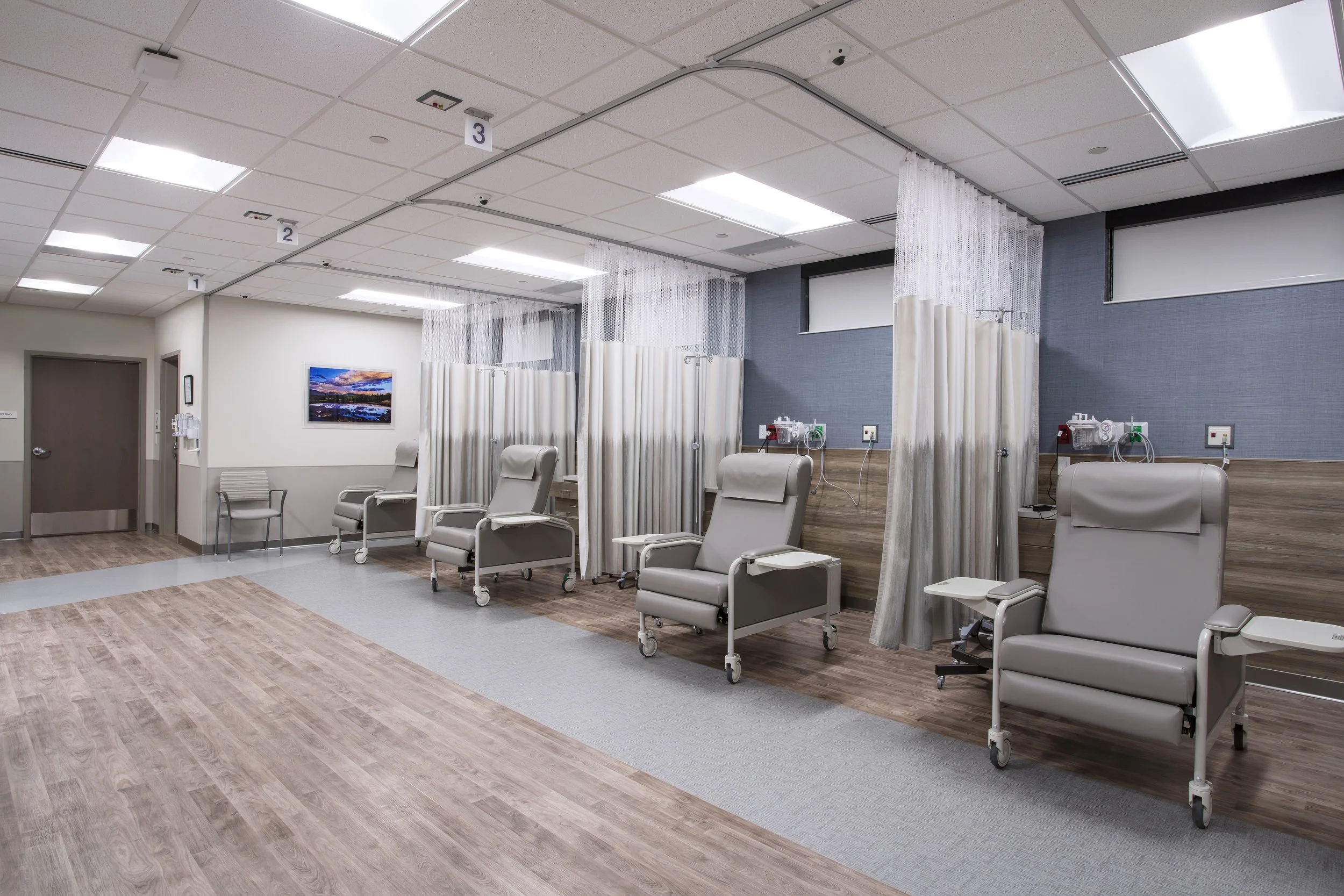 Hospital room with medical chairs, privacy curtains, and medical equipment on the walls, including a television, with ceiling lights and a wood and tile floor.