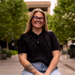 A woman with glasses, long hair, and tattoos smiling outdoors on a sidewalk lined with trees.
