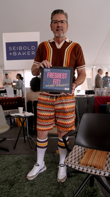 Man standing in a tent holding a sign that says "KDA Freshest Fit Award Winner." He is dressed in colorful, patterned shorts, a retro sweater, and knee-high socks with stripes.