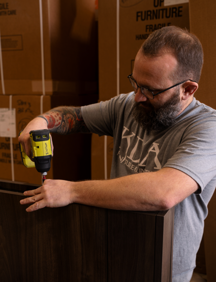 A man with a beard and glasses assembling furniture using a electric screw driver in a warehouse.