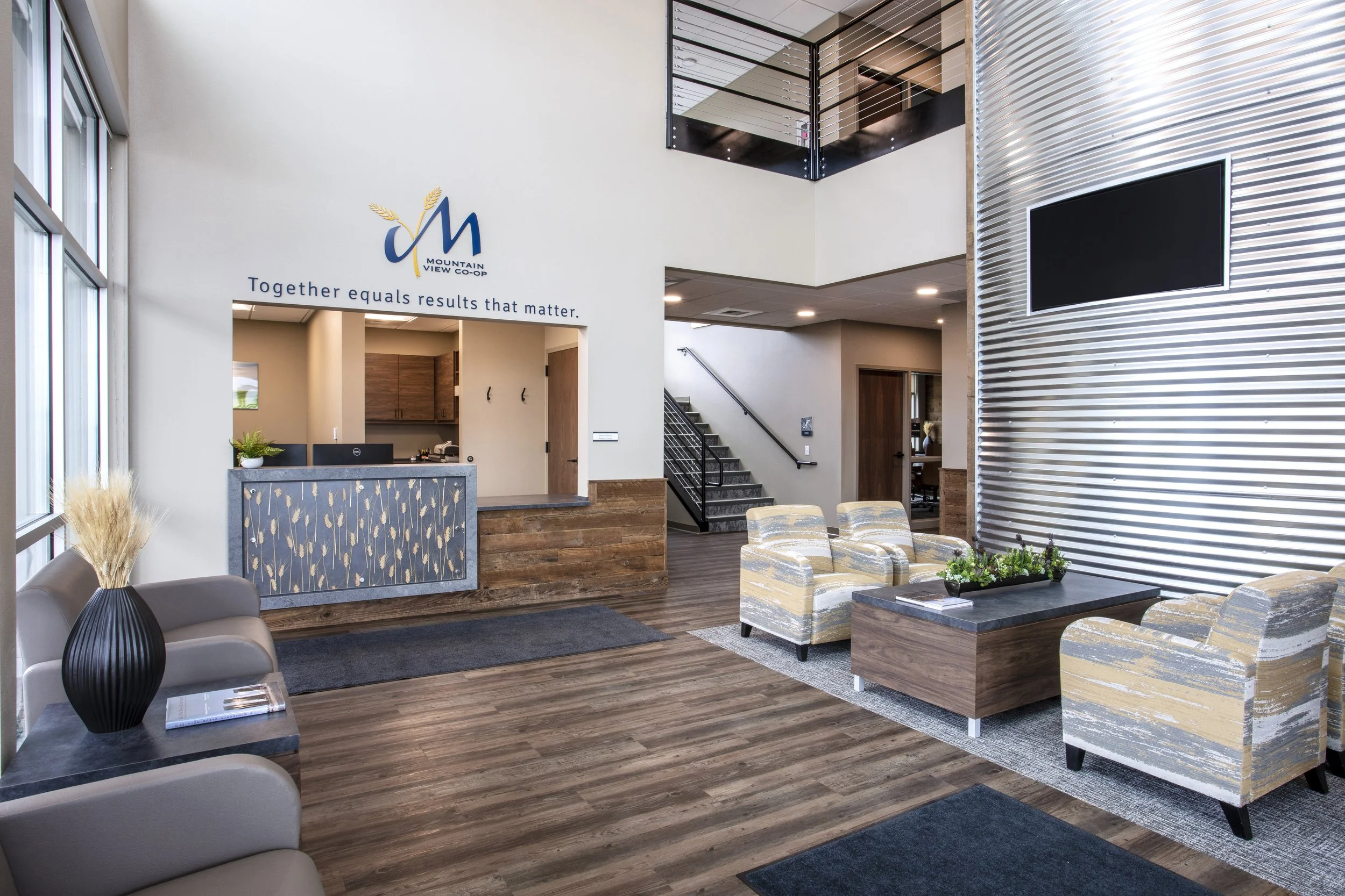 Interior of a modern office lobby with a reception desk, seating area with armchairs, a staircase, and a wall-mounted TV.