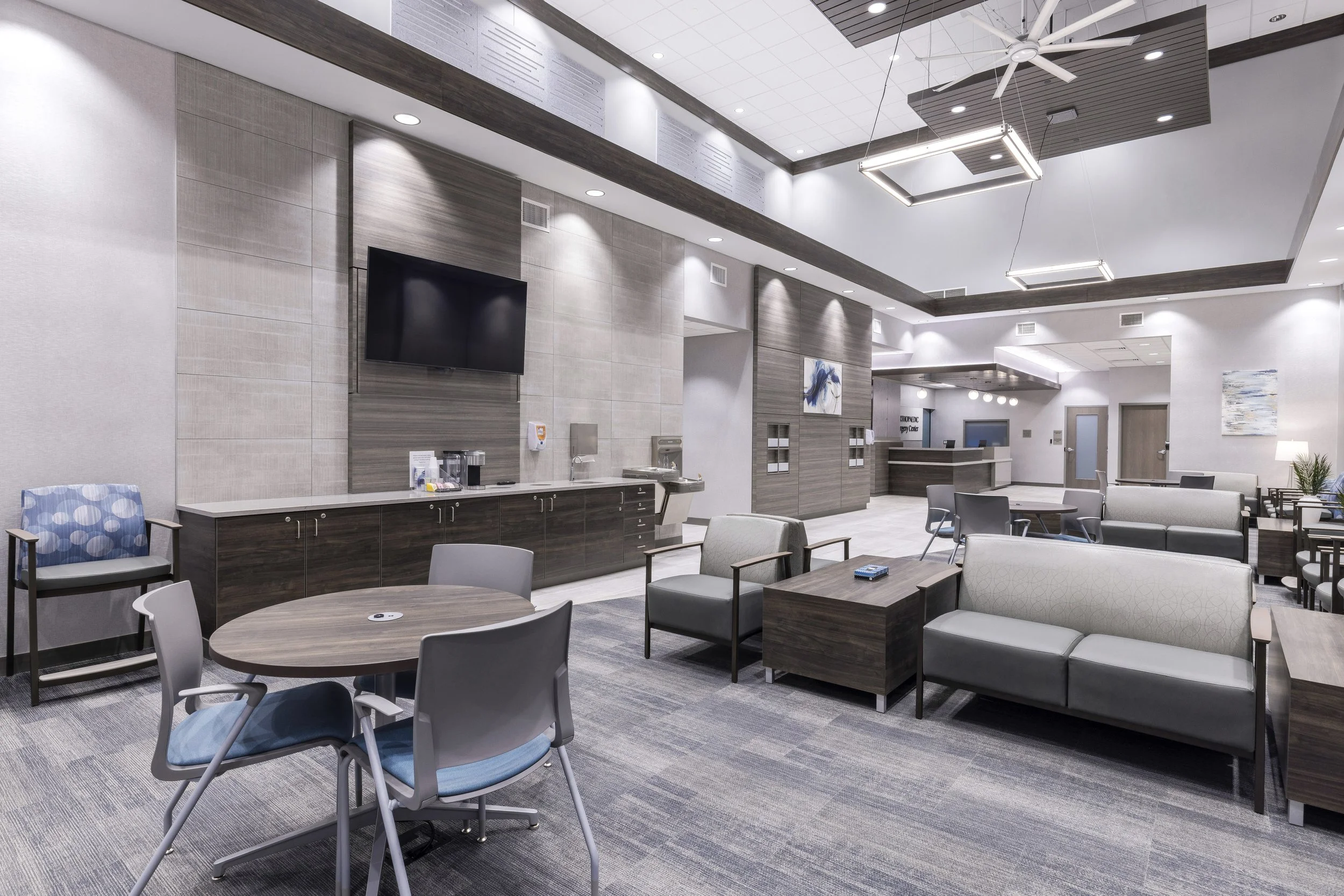 Modern waiting room with beige and gray color scheme, featuring chairs, tables, a large wall-mounted flat-screen TV, artwork on the walls, and ceiling fans.