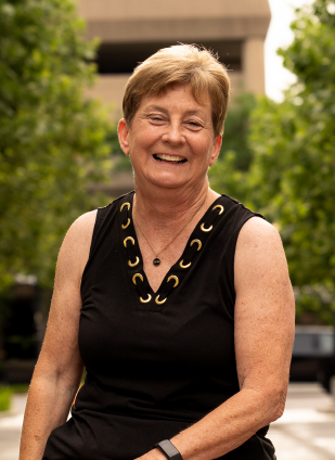 Smiling older woman with short hair wearing a black sleeveless top with a decorative neckline, outdoors with green trees in the background.