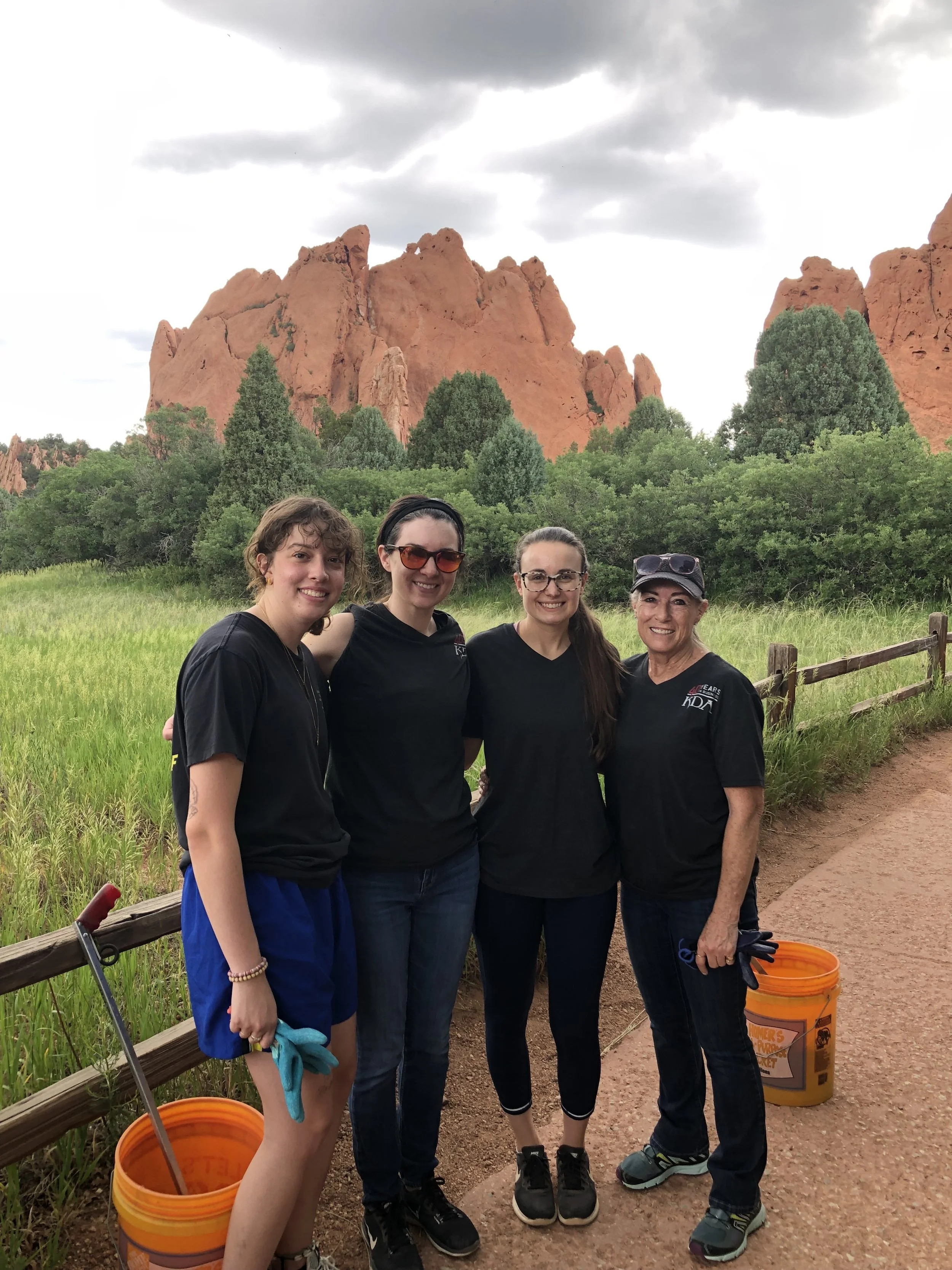 Four women standing outdoors in front of red rock formations and green trees, smiling at the camera during cloudy weather, with orange buckets nearby and a wooden fence on the side.