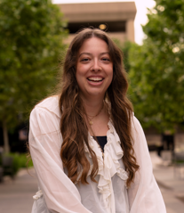 Young woman with long wavy brown hair smiling outdoors, wearing a white blouse, in a park-like setting.