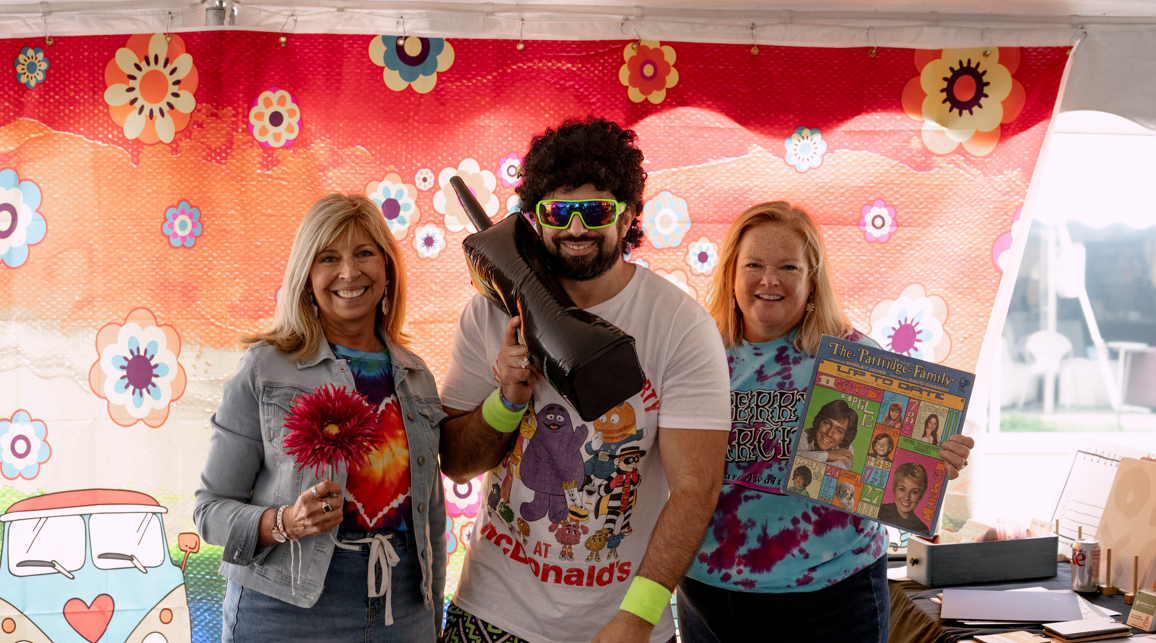Three people at a colorful outdoor party with a flower-patterned backdrop. The person in the middle wears sunglasses, a white T-shirt, and holds a black inflatable guitar. The woman on the left holds a pink flower, and the woman on the right holds a 