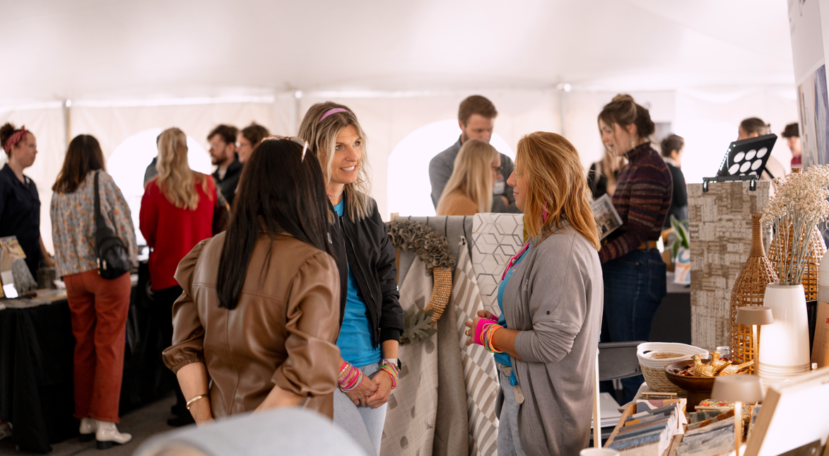 People at an indoor market or craft fair, chatting and browsing home decor items and textiles.