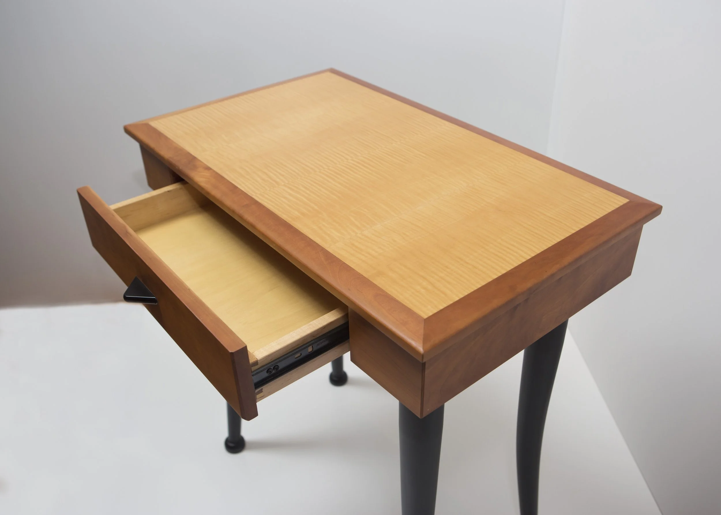 A wooden nightstand with one open drawer, showing a light-colored interior. The nightstand has a natural wood finish on the top surface and darker wood framing, with black legs supporting it.