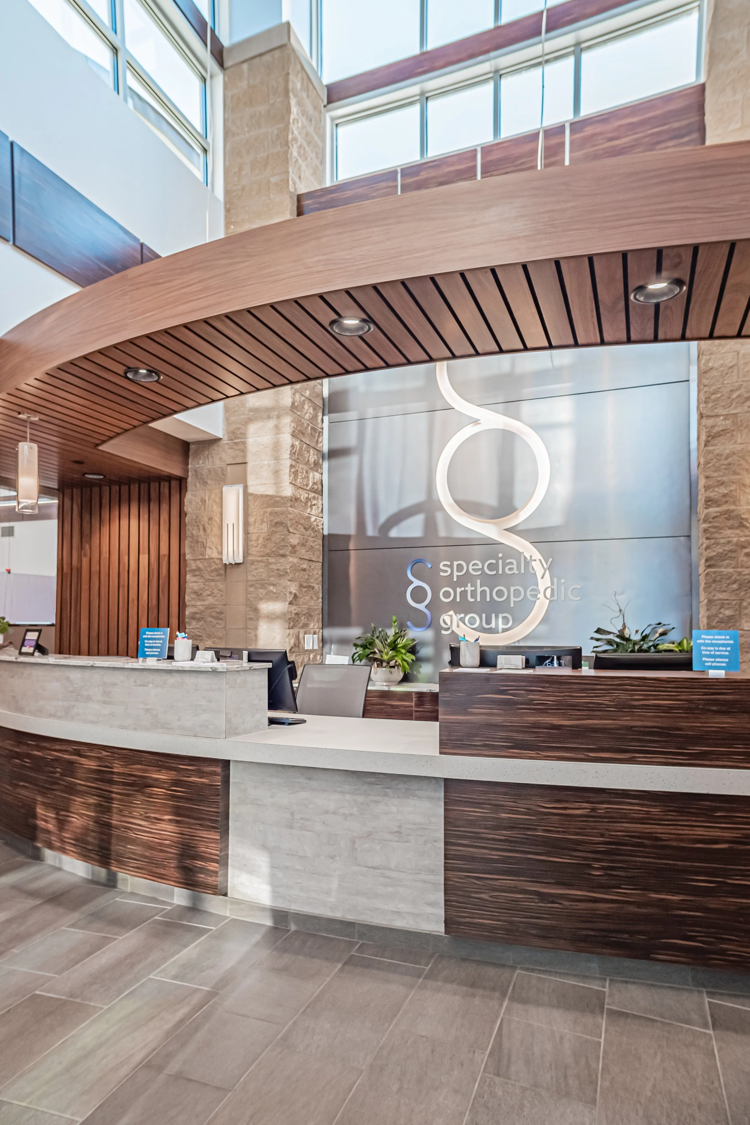 Reception desk at a medical office with wood accents, plants, and a sign reading 'specialty orthopedic group' in a modern, well-lit lobby with large windows.