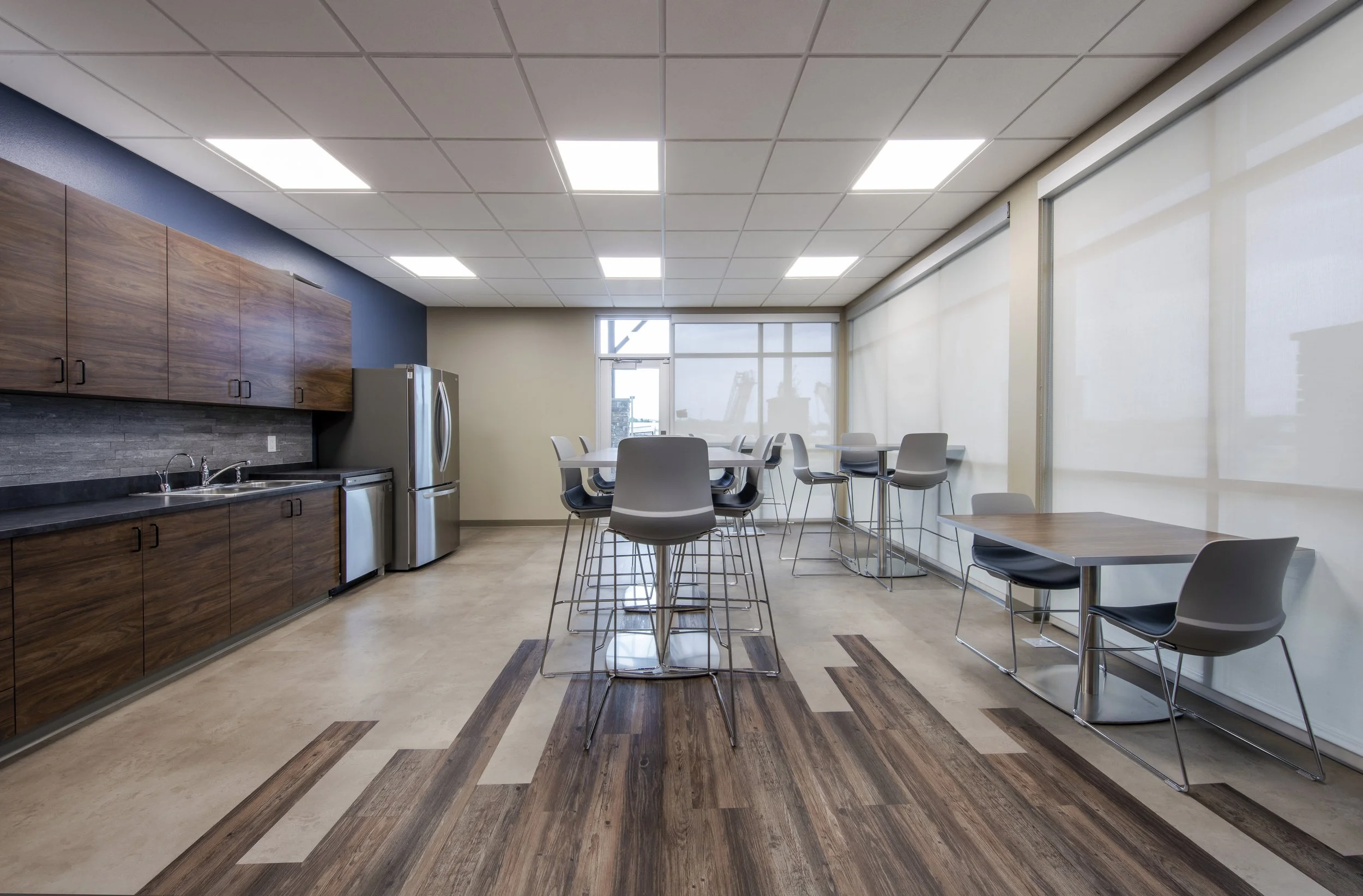 Empty modern office break room with tables and chairs, kitchen area with wooden cabinets, stainless steel refrigerator, and large windows with blinds.
