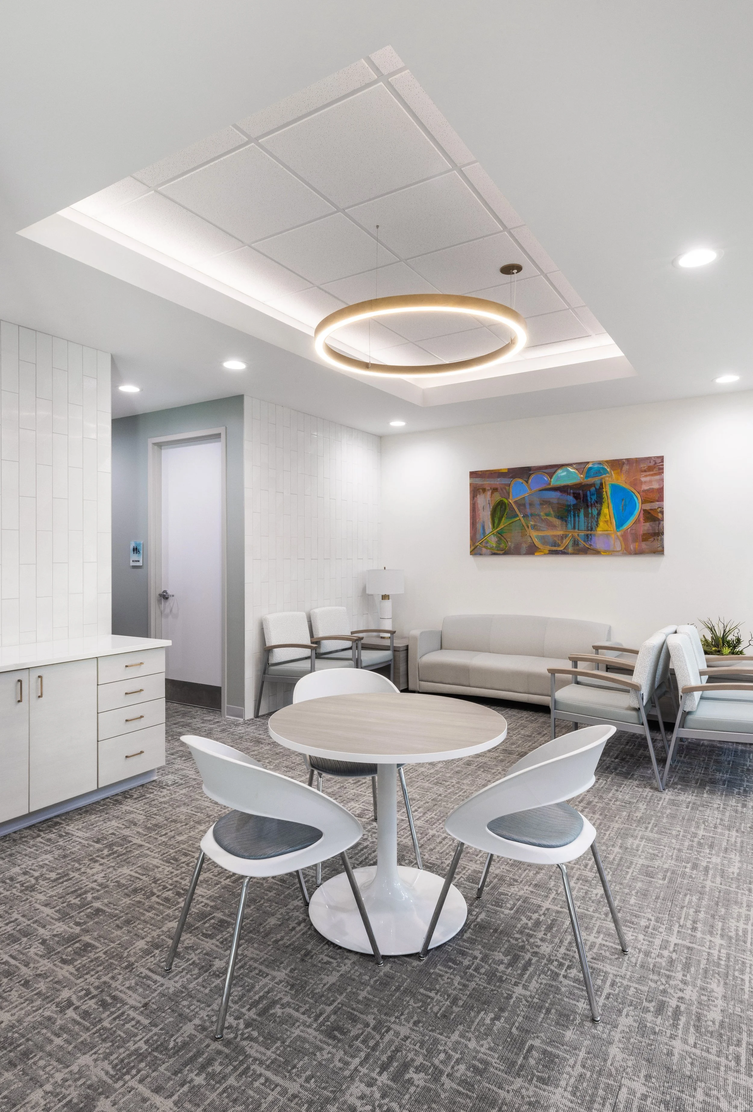 A modern waiting room or lounge area with a round white table and four white chairs with gray cushions, a white sofa, a small side table with a white lamp, and an abstract colorful painting on the wall. The ceiling has recessed lighting and a circular modern light fixture, with a gray patterned carpet on the floor.