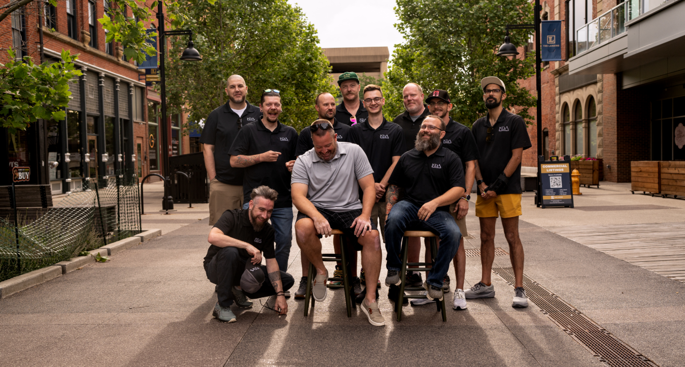 A group of eleven men gathered on an outdoor urban street, some sitting on stools and others standing behind, all smiling and dressed casually in black and gray shirts, with trees and storefronts in the background.