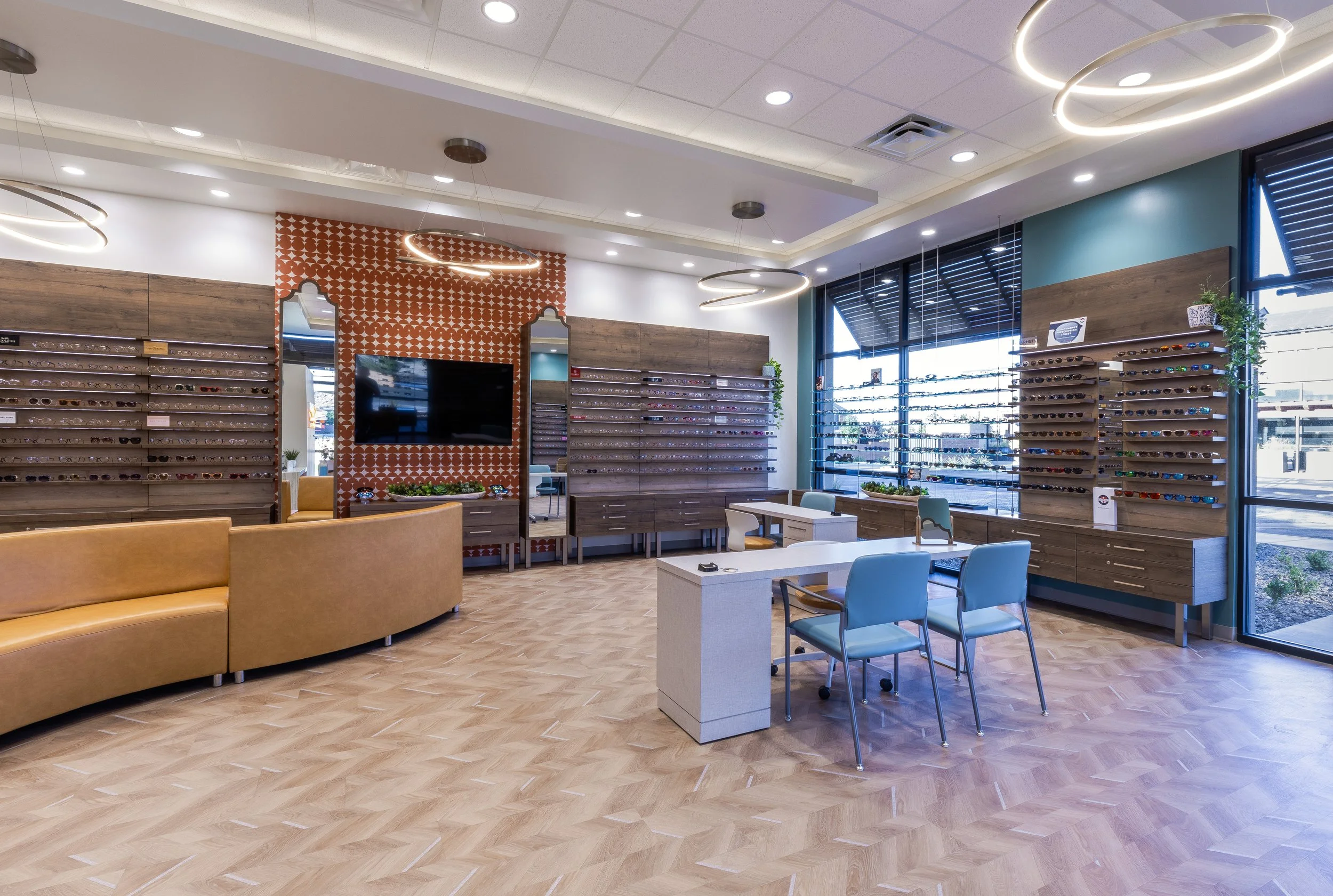 Interior of an eyeglass store with wooden display shelves filled with various eyeglasses, a large flat screen TV, modern circular ceiling lights, and ample sunlight coming through large windows.