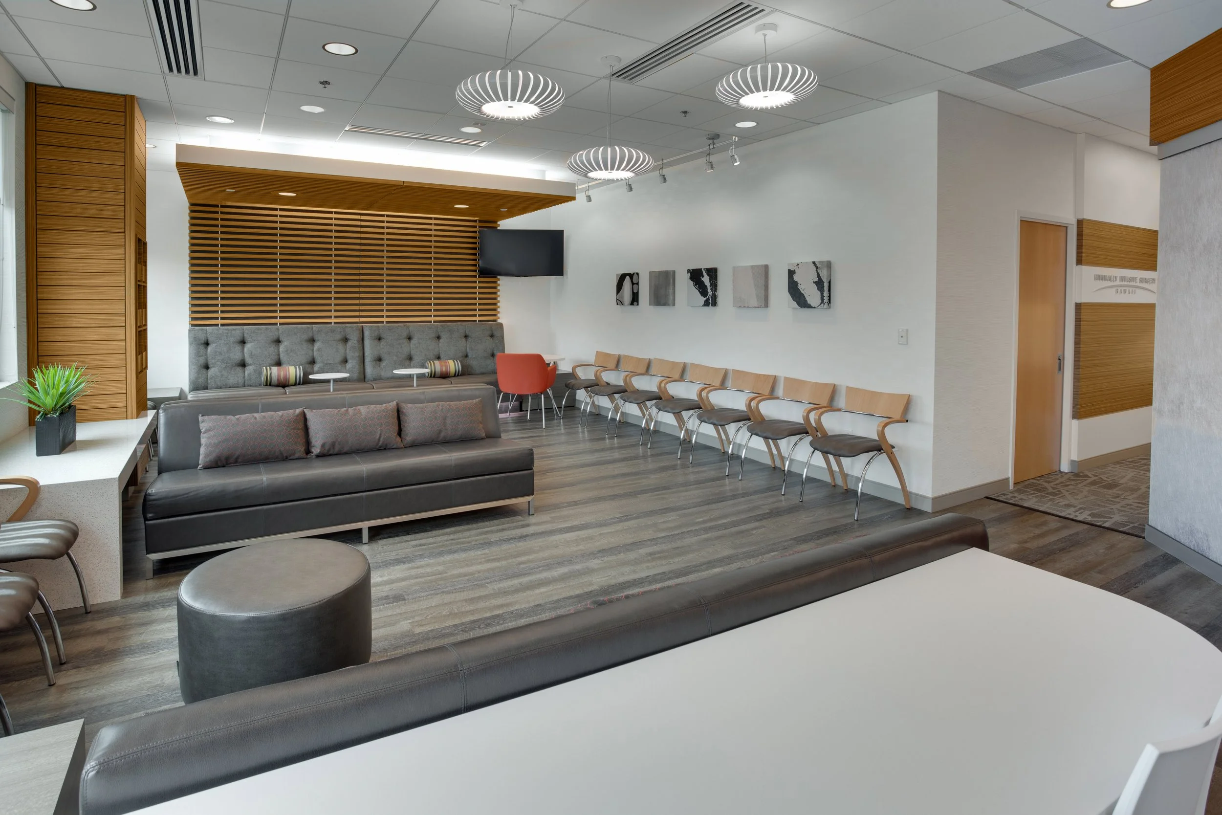 Interior of a modern waiting room with gray and wooden furniture, a row of chairs along the wall, pendant lights, and artwork on the walls.