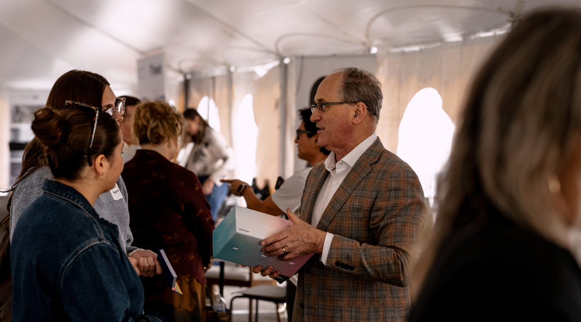 A man in glasses and a plaid blazer talking to a group of women at an indoor event, holding a large book or folder.
