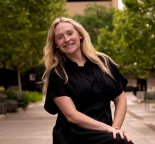 A young woman with long blonde hair smiling outdoors in a park-like setting with trees and buildings in the background.