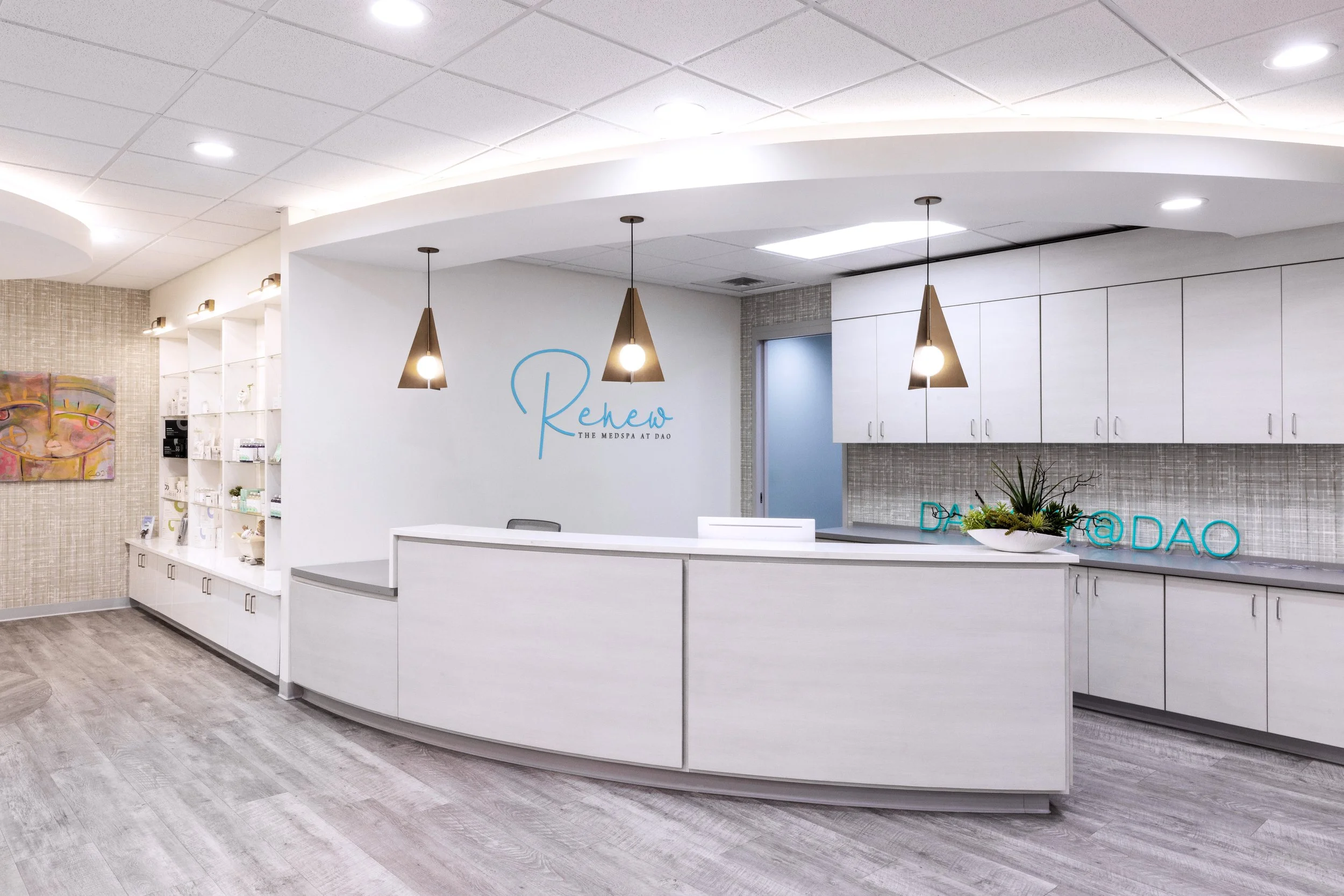 Interior of a medical spa with a white reception desk, modern hanging pendant lights, white cabinetry, decorative plant, and artwork on the wall.