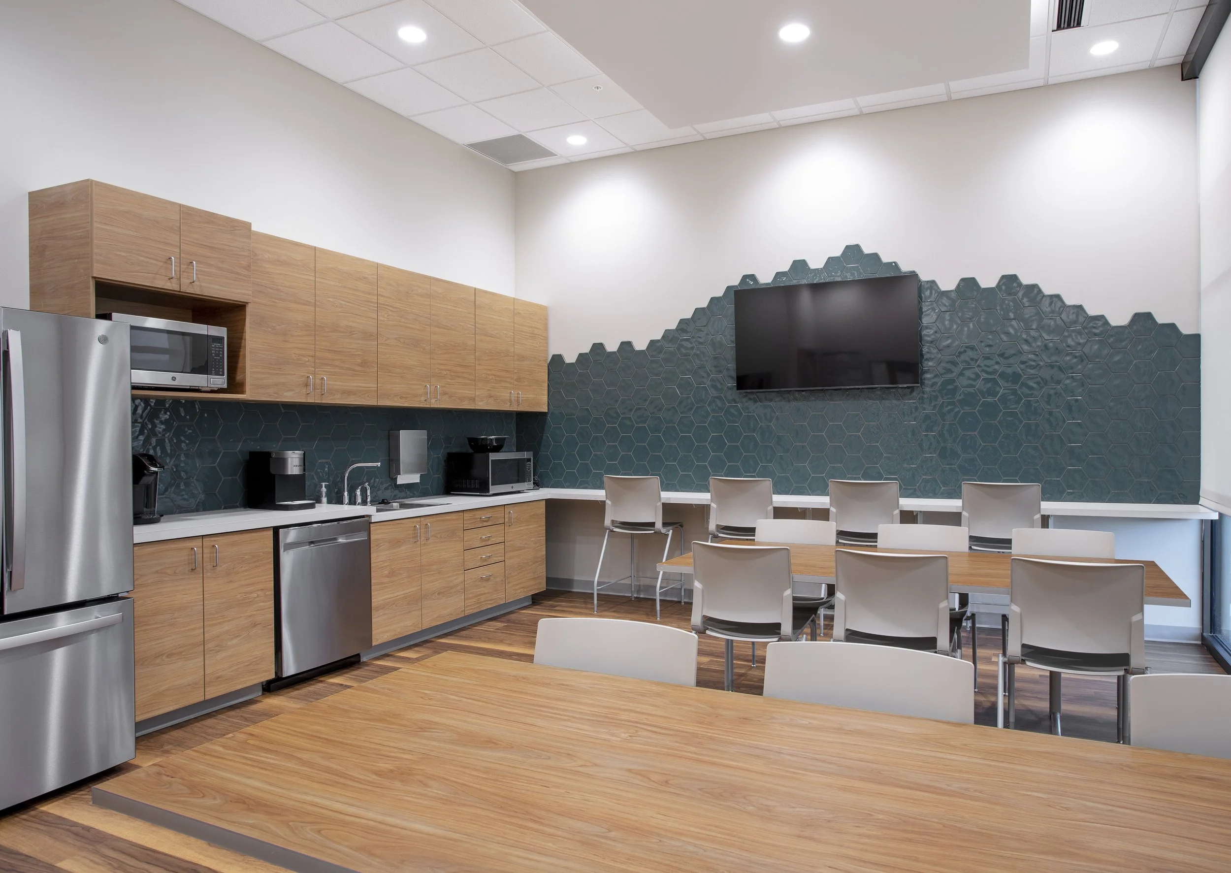 A modern kitchen and dining area with wooden cabinets, a microwave, stainless steel refrigerator, and a black hexagonal tile backsplash. There are white chairs around wooden tables, and a large flat-screen TV mounted on the wall.
