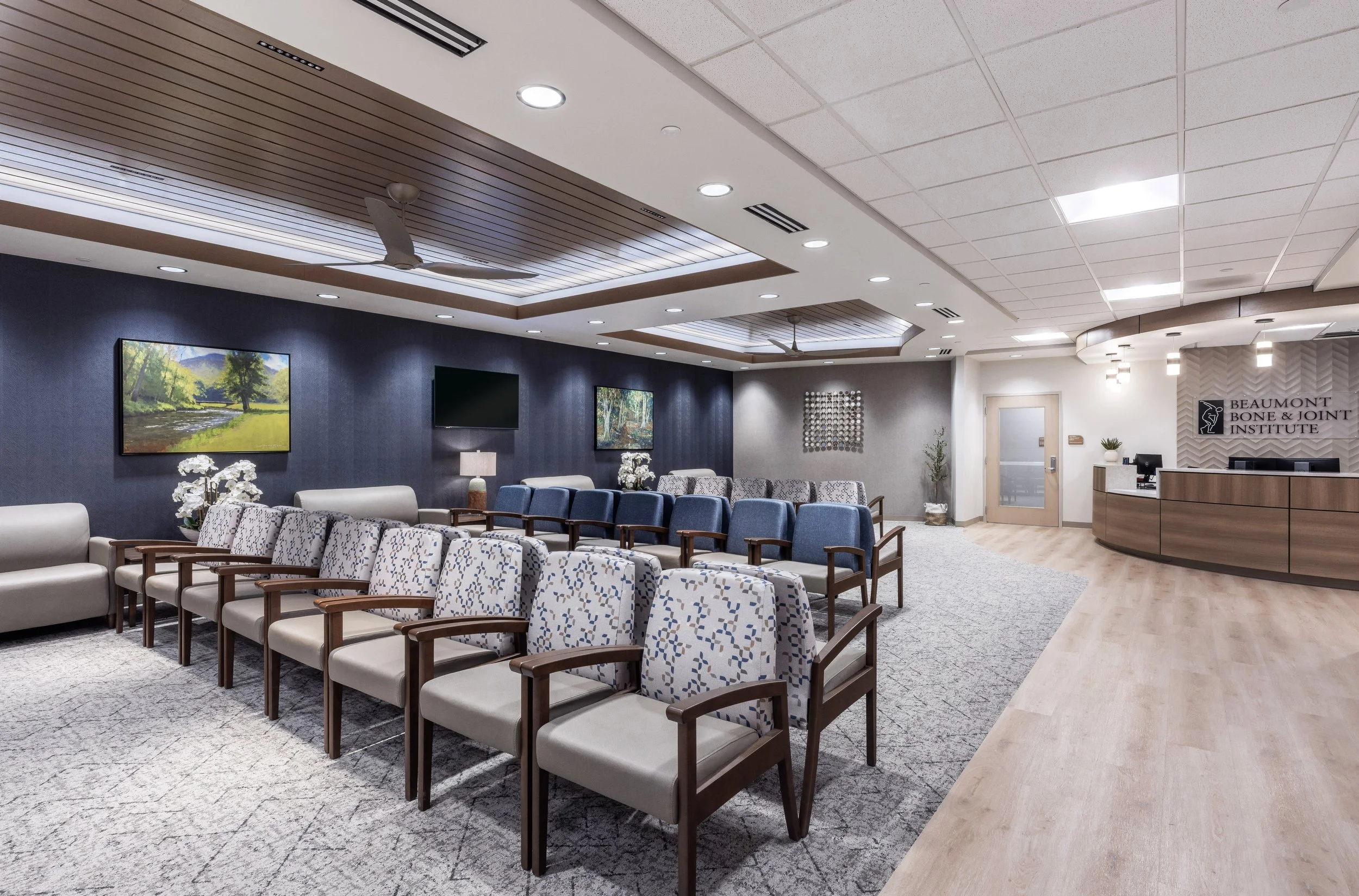A waiting area with multiple rows of beige and blue seats, dark blue wall with two landscape paintings, a flat screen TV, and a reception desk with a sign for Beaumont Bone & Joint Institute.