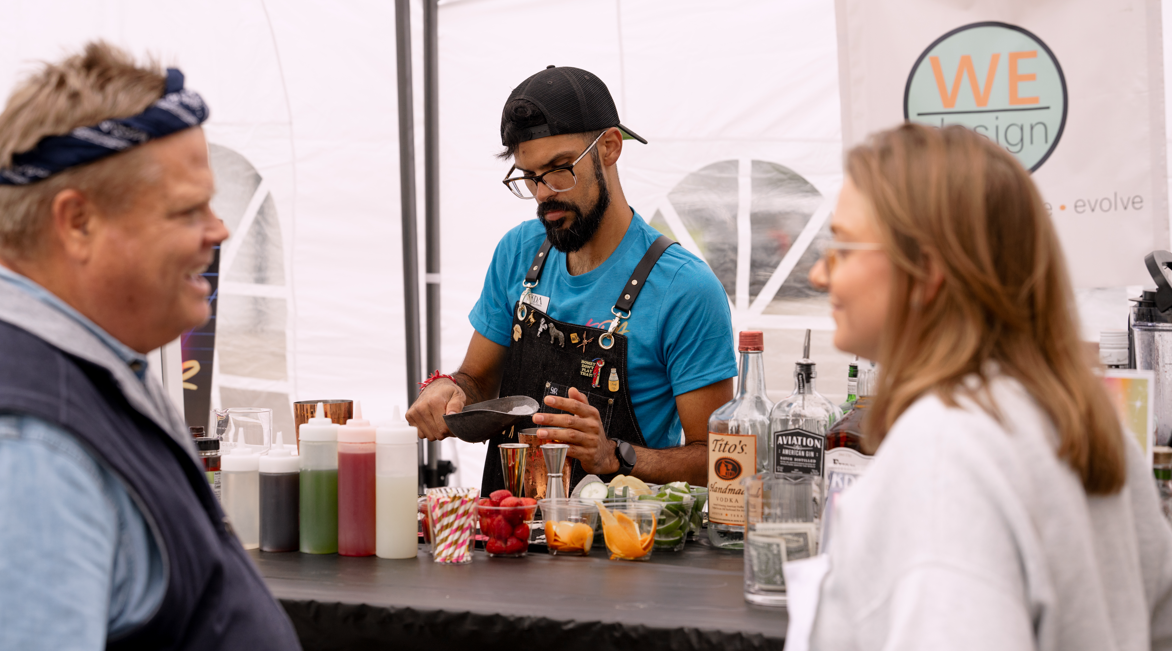 A bartender preparing drinks at a bar counter with colorful ingredients, while two customers, a man and a woman, watch and engage in conversation.