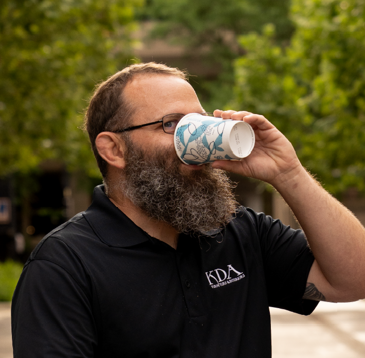 A bearded man wearing glasses and a black polo shirt with 'KDA' embroidered on it drinks from a paper cup outdoors with green trees in the background.