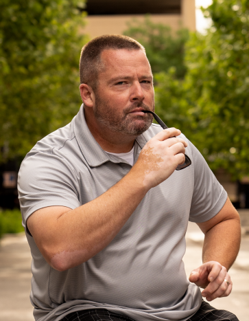 A man with a beard and short hair, wearing a gray polo shirt, holding a pipe to his lips, outdoors with green trees and buildings in the background.