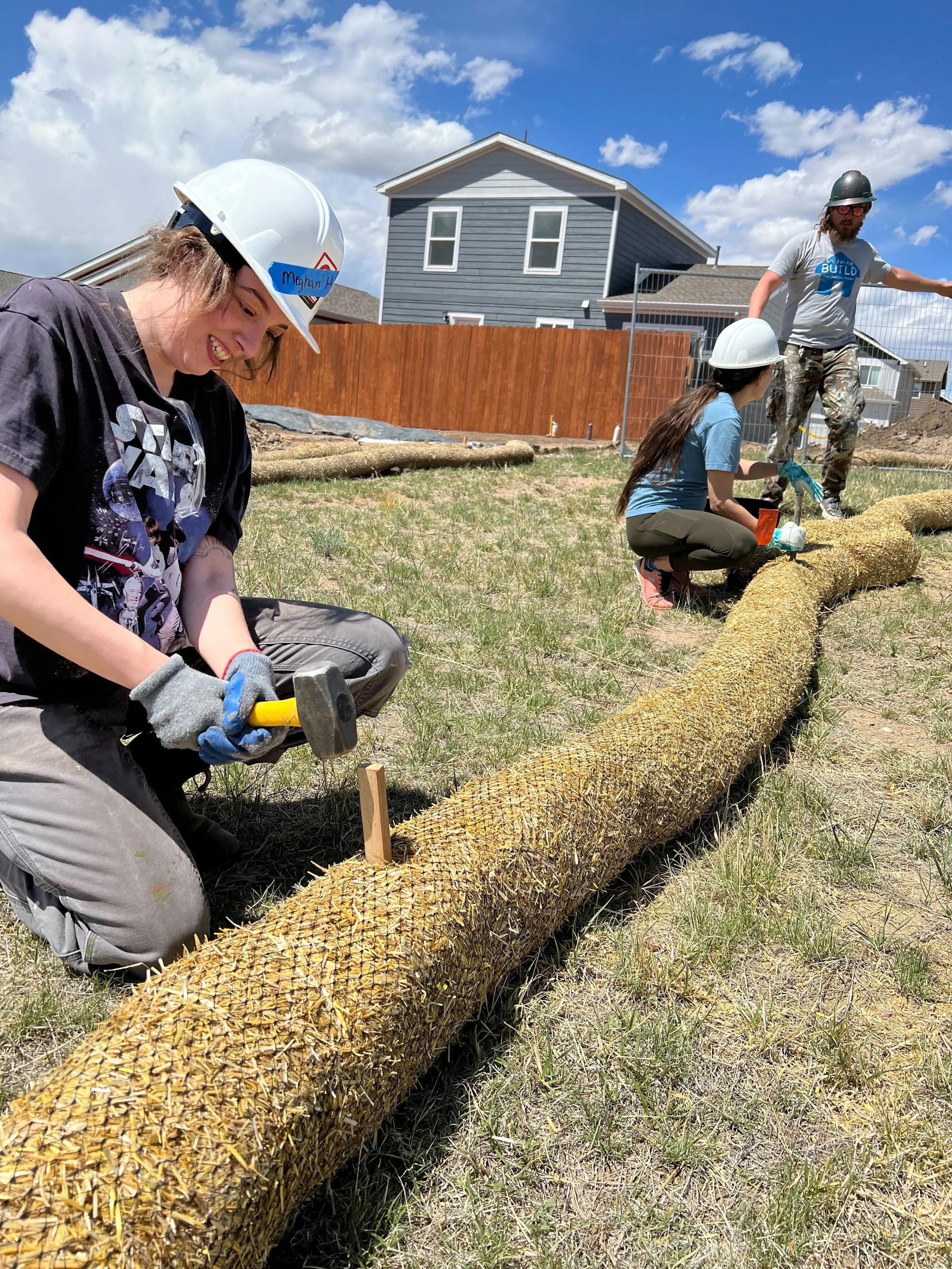 Three people working on a construction project outdoors, wearing safety helmets, with a large rolled-up natural fiber mat, a wooden stick, and tools on the ground, in front of suburban houses and a wooden fence under a partly cloudy sky.