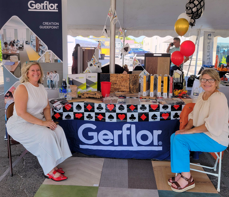 Two women sitting at a Gerflor booth with balloons and promotional items, inside a tent at an event.