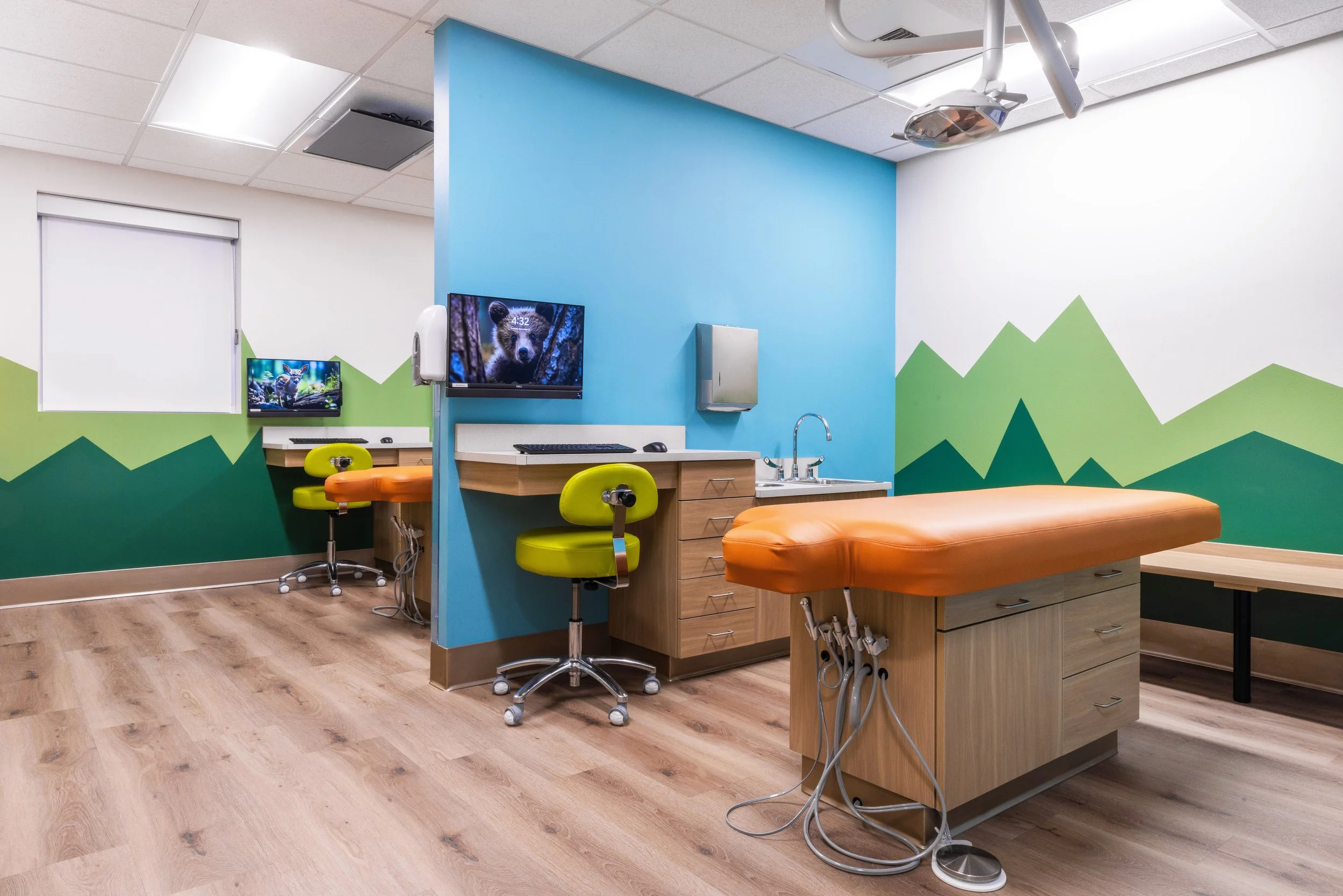 Medical examination room with orange exam table, wooden cabinets, green chairs, and a social media screen showing a bear.