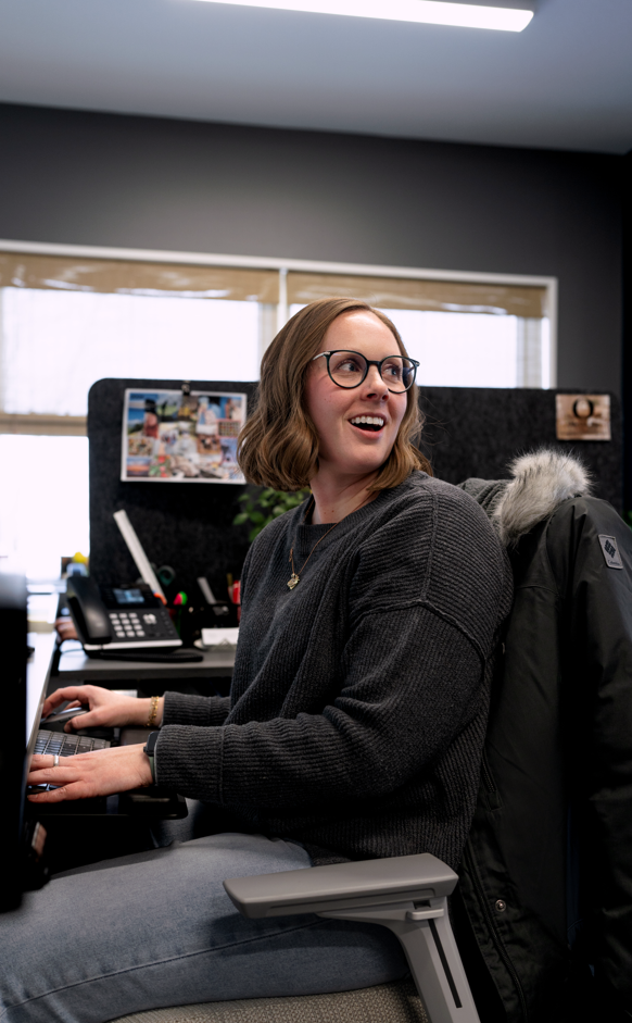 A young woman with glasses and shoulder-length brown hair working at a desk in an office, smiling and looking slightly to her right.