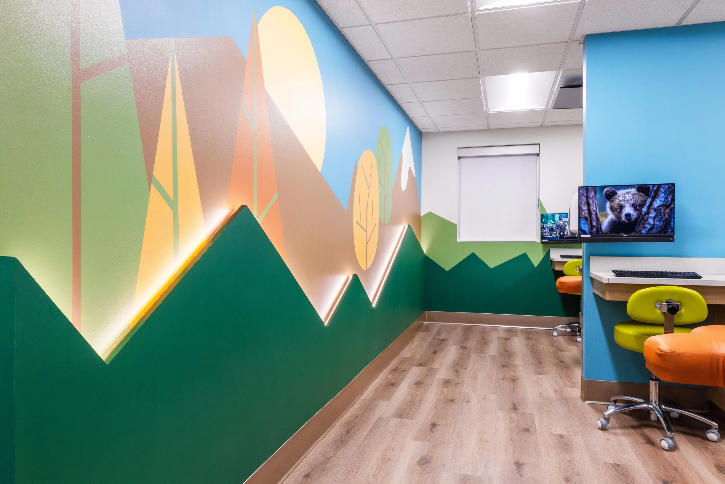 Colorful children's playroom or classroom with mountain and forest wall mural, bright orange chairs, computer desks, a mounted monitor showing a bear, and a window with a white blind.