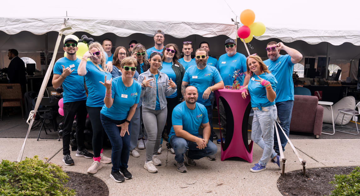 Group of people celebrating under a white tent, wearing matching blue shirts, with some holding colorful balloons, gathered around a pink table with small gifts or decorations, smiling and making gestures.