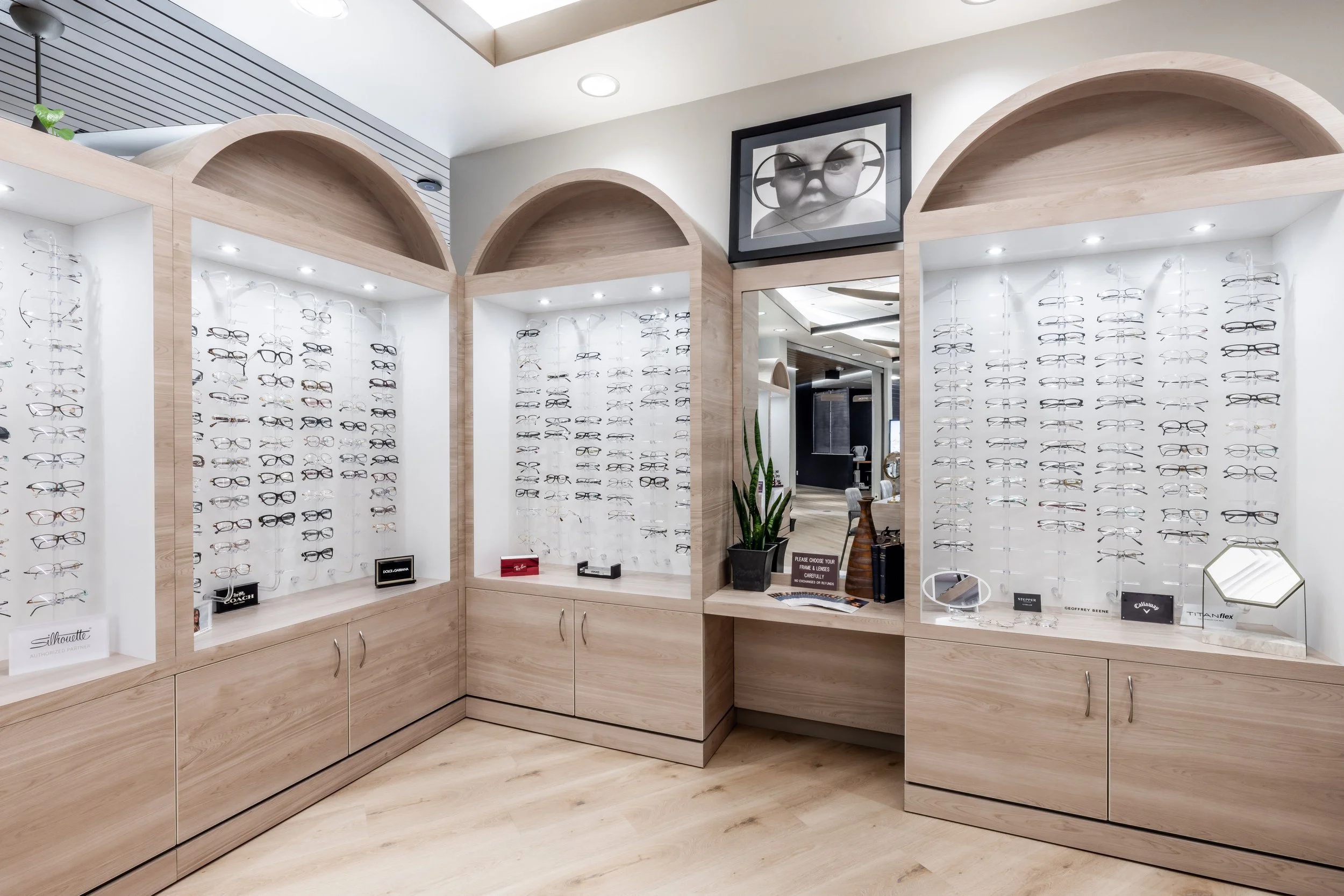 Eyewear display in a store showcasing various eyeglasses on white walls with wooden framing and cabinets below. A framed photo of a child with glasses decorates the wall.