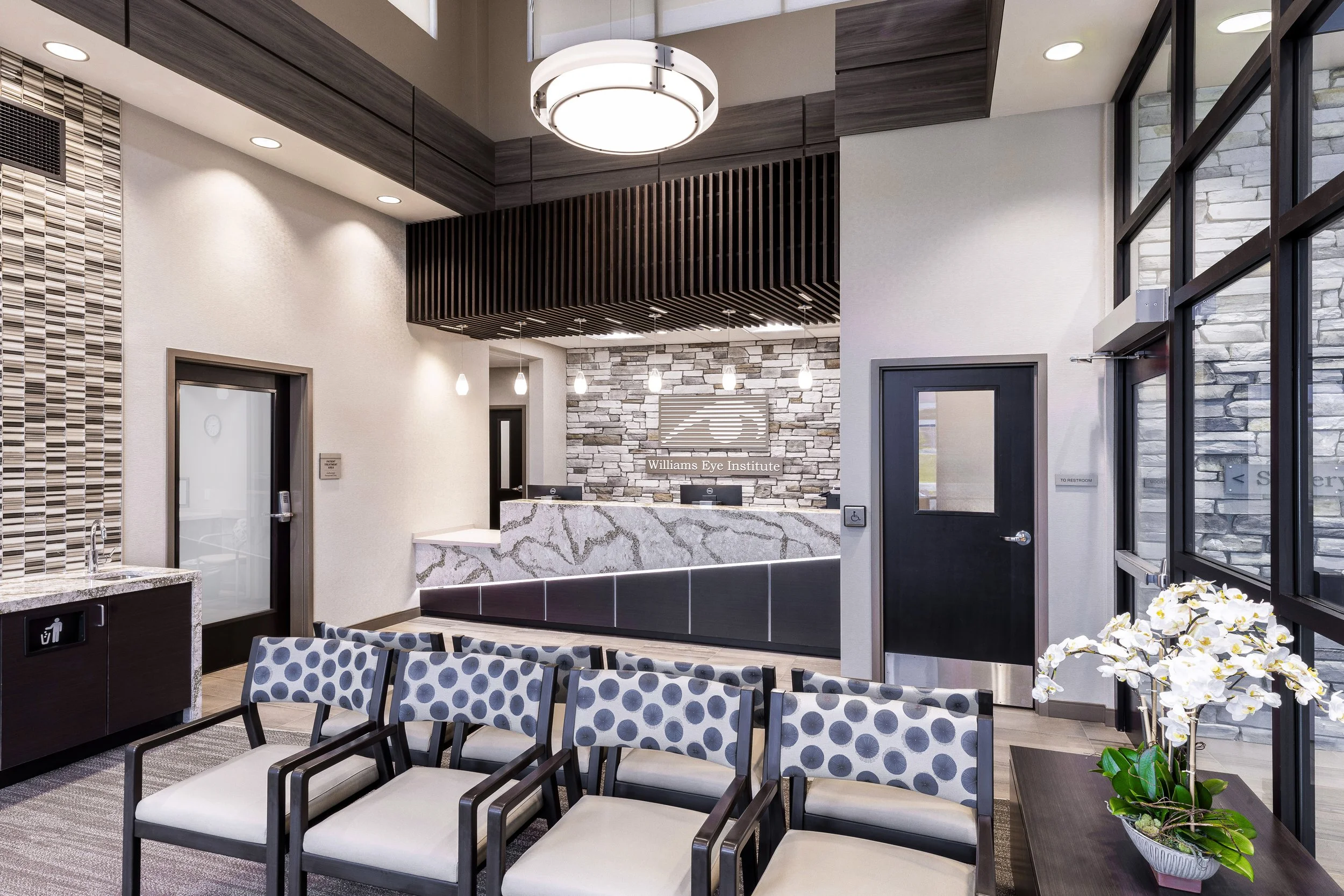 Reception area of the Willams Eye Institute with a marble counter, a stone accent wall, modern light fixtures, and seating with polka dot patterned cushions.