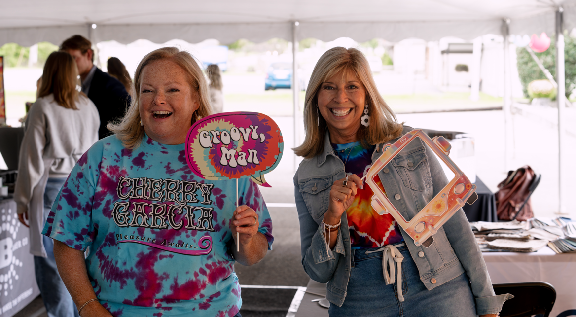 Two women smiling at a daytime outdoor event, one holding a sign that says "groovy man" and the other holding a toy camera frame, under a tent with tables and other people in the background.