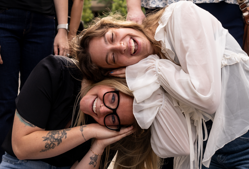 Two women smiling and leaning their heads together in an outdoor setting, appearing happy and friends.