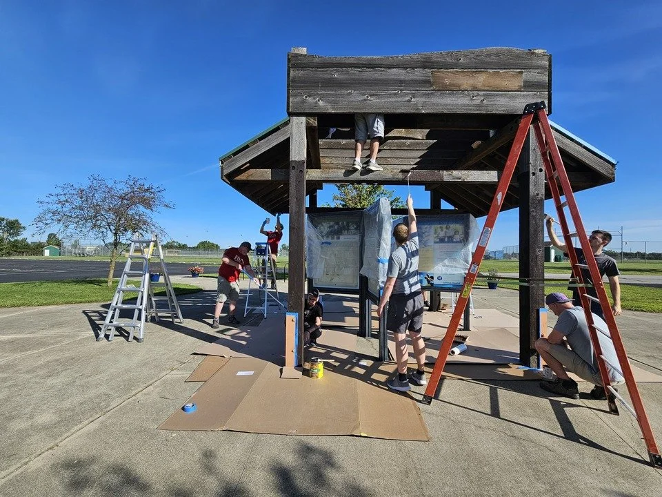 Group of people assembling and repainting a playground structure outdoors on a sunny day, with ladders and paint supplies.