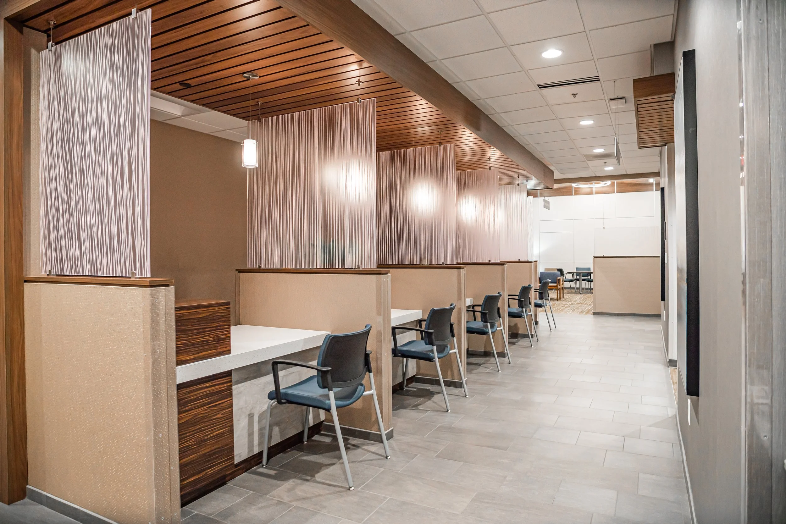Interior of a waiting area in a modern building with chairs, wooden accents, suspended light fixtures, and partial partition panels.