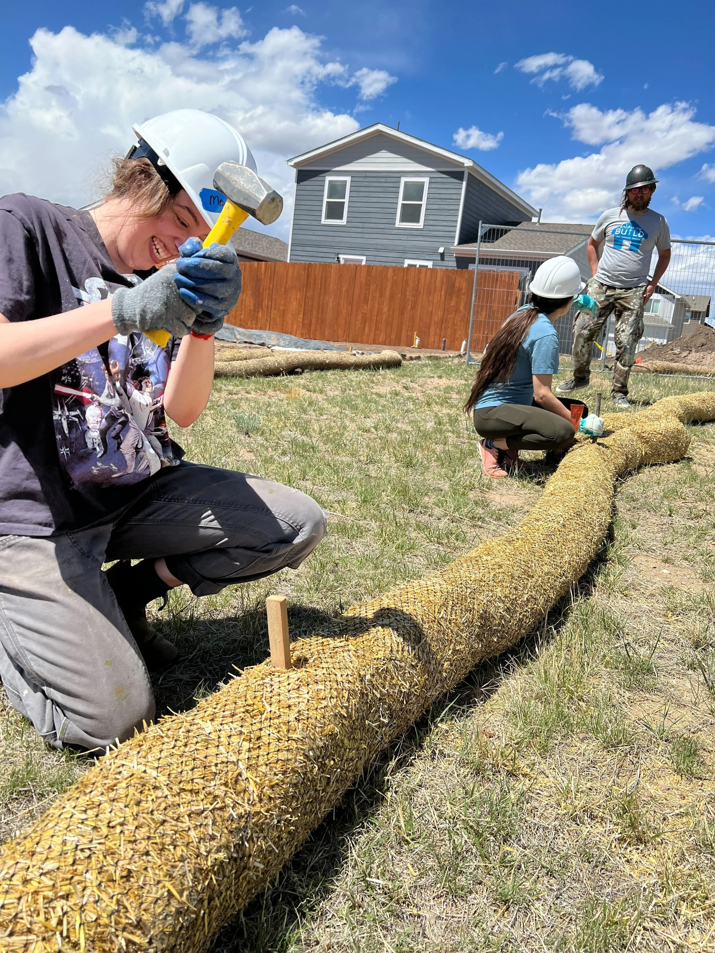 Three people wearing safety helmets working with a large, cut tree trunk outdoors on a sunny day, with houses and a blue sky in the background.