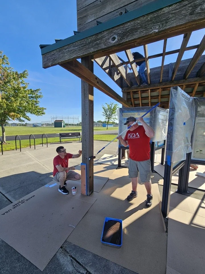 Three people working on building or repairing a wooden structure outdoors on a sunny day. One person is on the upper level, working with tools, while two others are on the ground, painting or inspecting the lower part.