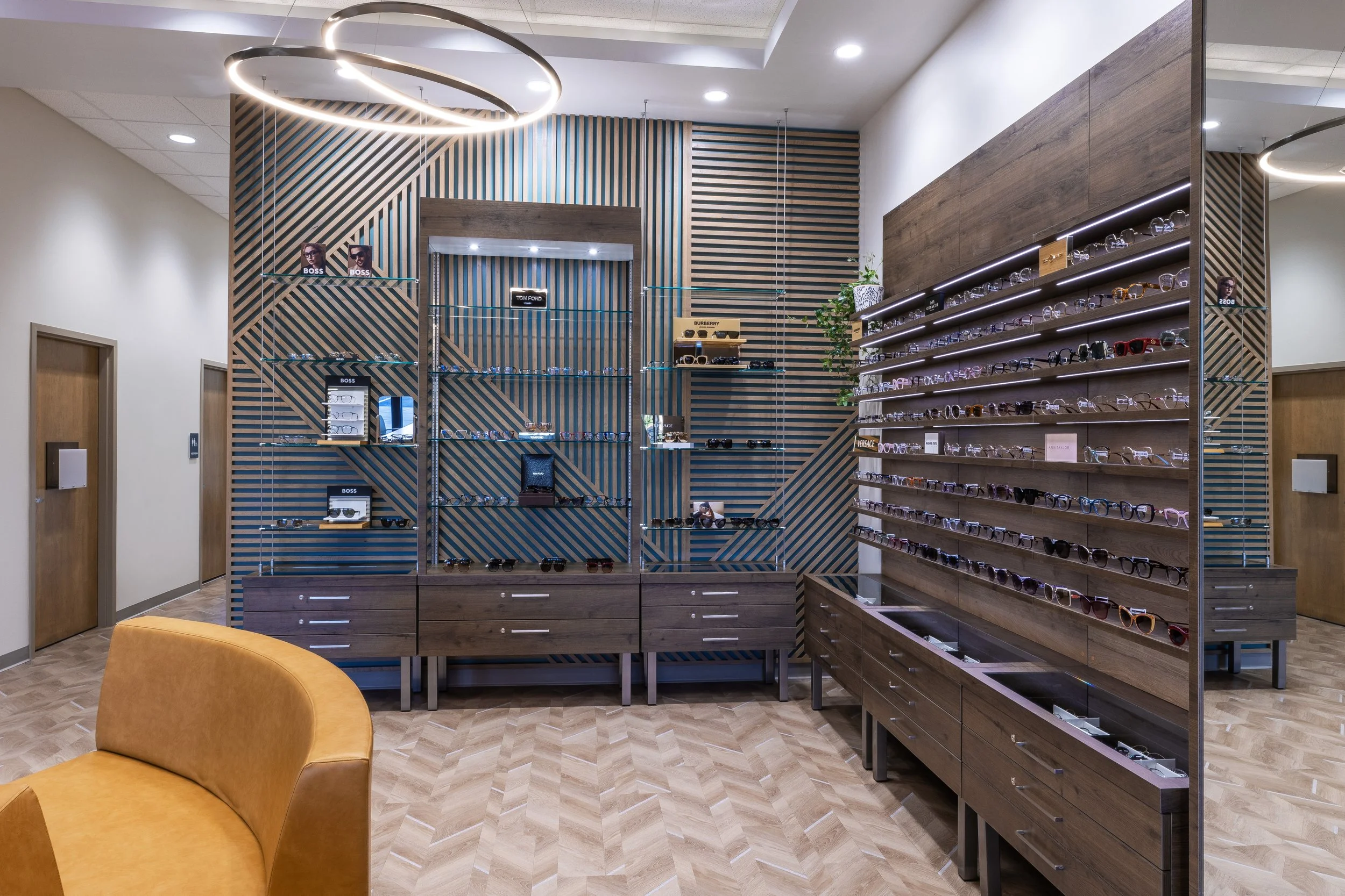 Interior of an eyewear store with wooden shelves displaying sunglasses and eyeglasses, modern circular ceiling lights, and a mustard-colored chair in the foreground.