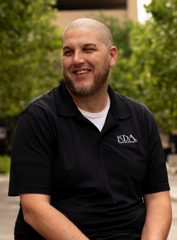 Man with a shaved head smiling outdoors, wearing a black polo shirt with a logo, in front of trees.