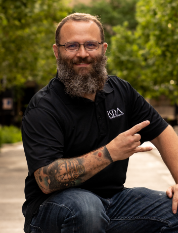A smiling man with glasses and a beard, wearing a black polo shirt with a logo, sitting outdoors on a bench or similar surface, pointing with his right hand.